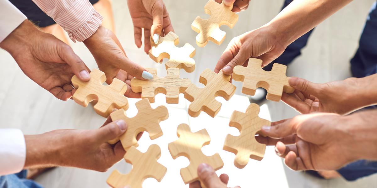 Several people holding wooden puzzle pieces together over a table
